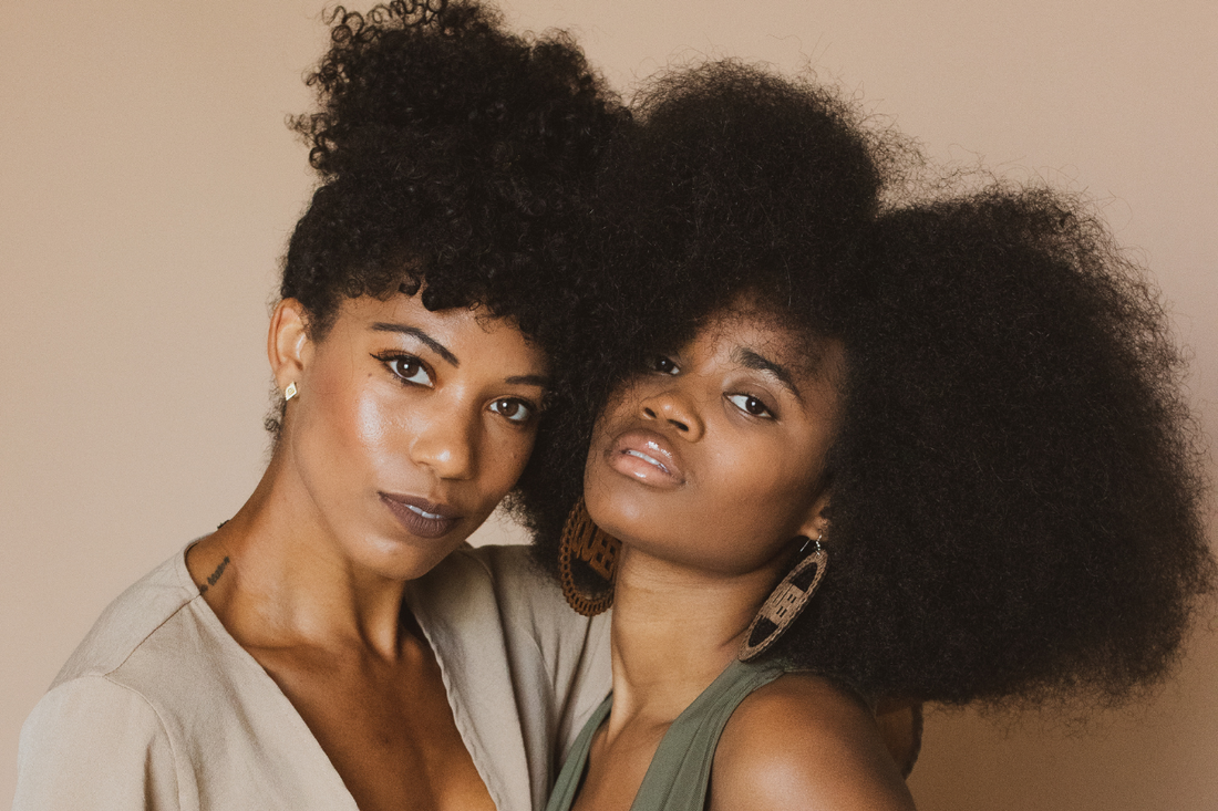 Two women with natural textured hair posing closely together, one with a curly updo and the other with a voluminous afro, against a neutral beige background.          Ask ChatGPT