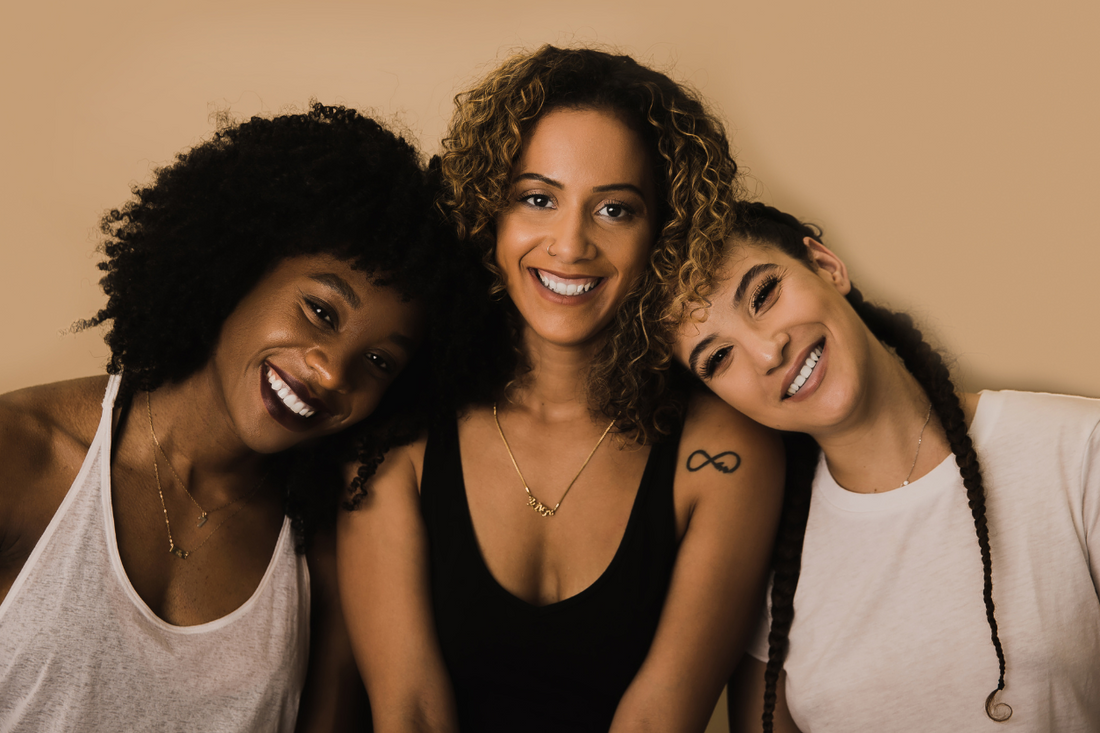Three women with different hair textures—kinky, curly, and wavy—smiling together against a tan background, celebrating hair diversity and joy.
