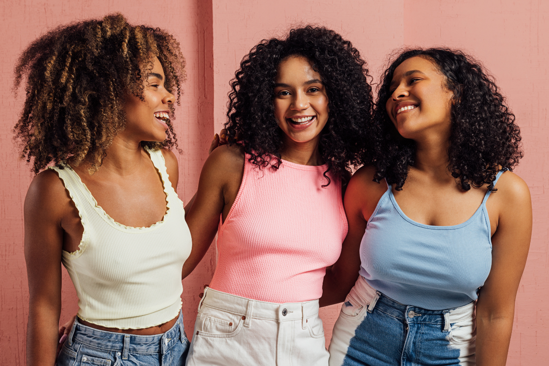 Three women with different curly hair textures smiling together against a pink background, showcasing natural curl pattern diversity.