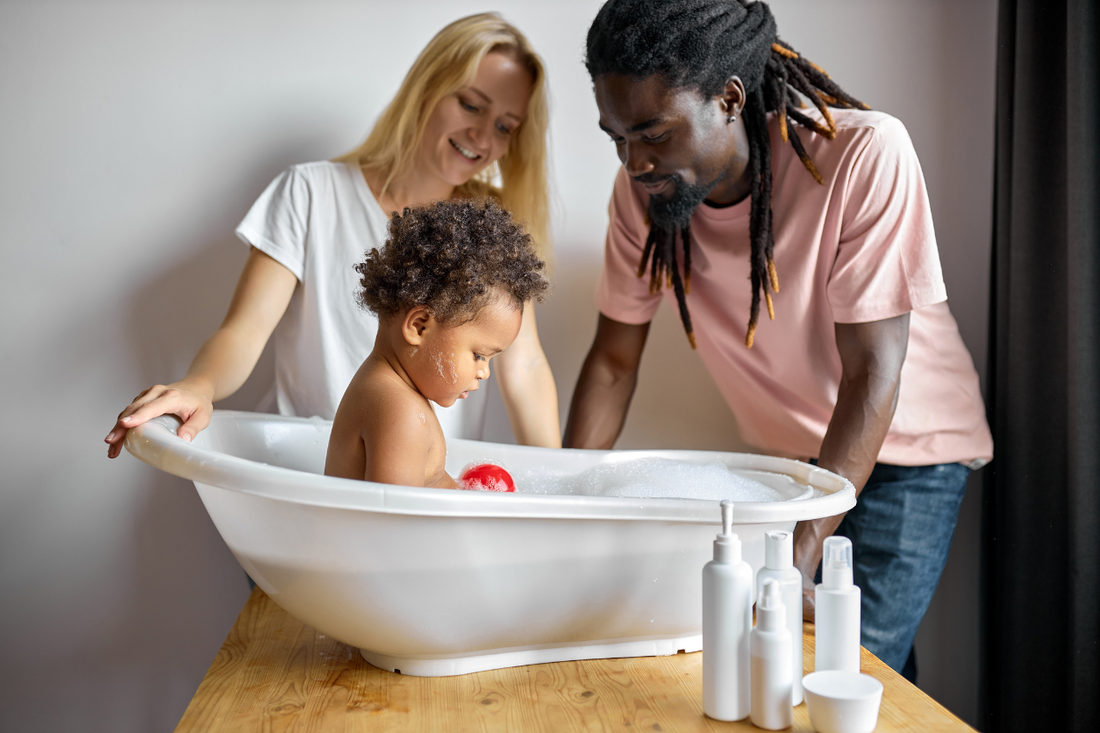 Parents bathing their toddler in a small tub with bubbles, surrounded by hair and skincare bottles.