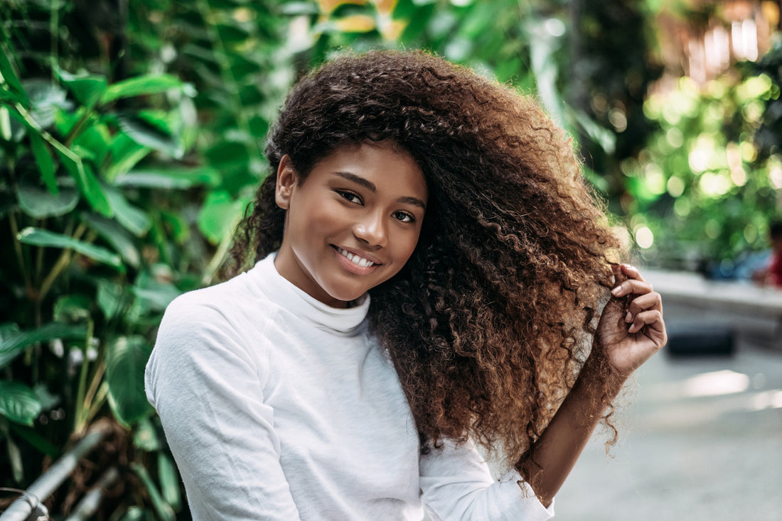 Smiling woman in white outside while holding her brown curly hair