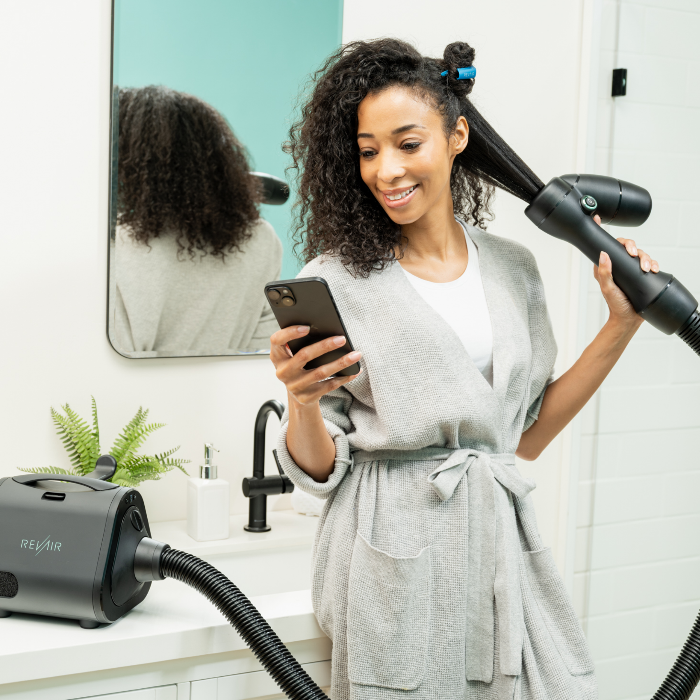 Woman in bathrobe scrolling on her phone with one hand while using the RevAir dryer with her other hand, showing one-handed operation.