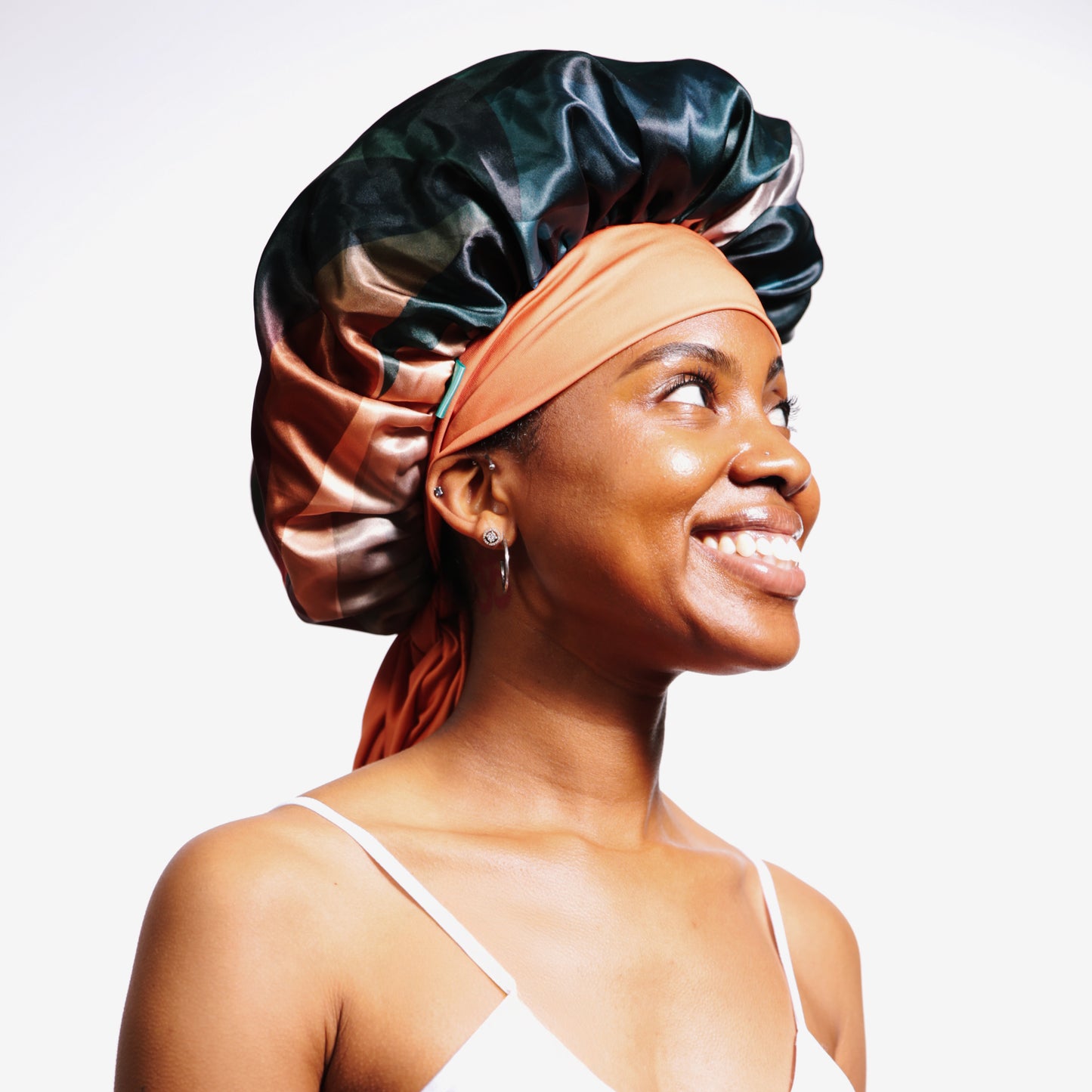 A smiling woman wearing a colorful satin bonnet with shades of teals, copper, and beige, with an orange head wrap tied around her head. She is dressed in a white spaghetti strap top, looking off to the side against a plain white background.