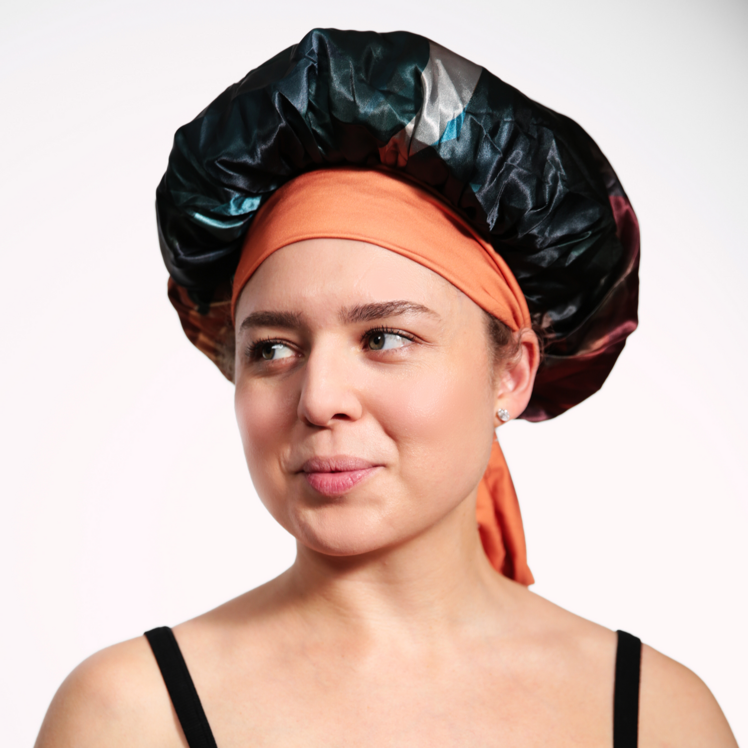 A woman wearing a colorful satin bonnet with shades of black, teal, and burgundy, paired with an orange head wrap tied around her head. She is dressed in a black spaghetti strap top, looking slightly to the side with a subtle smile, against a plain white background.