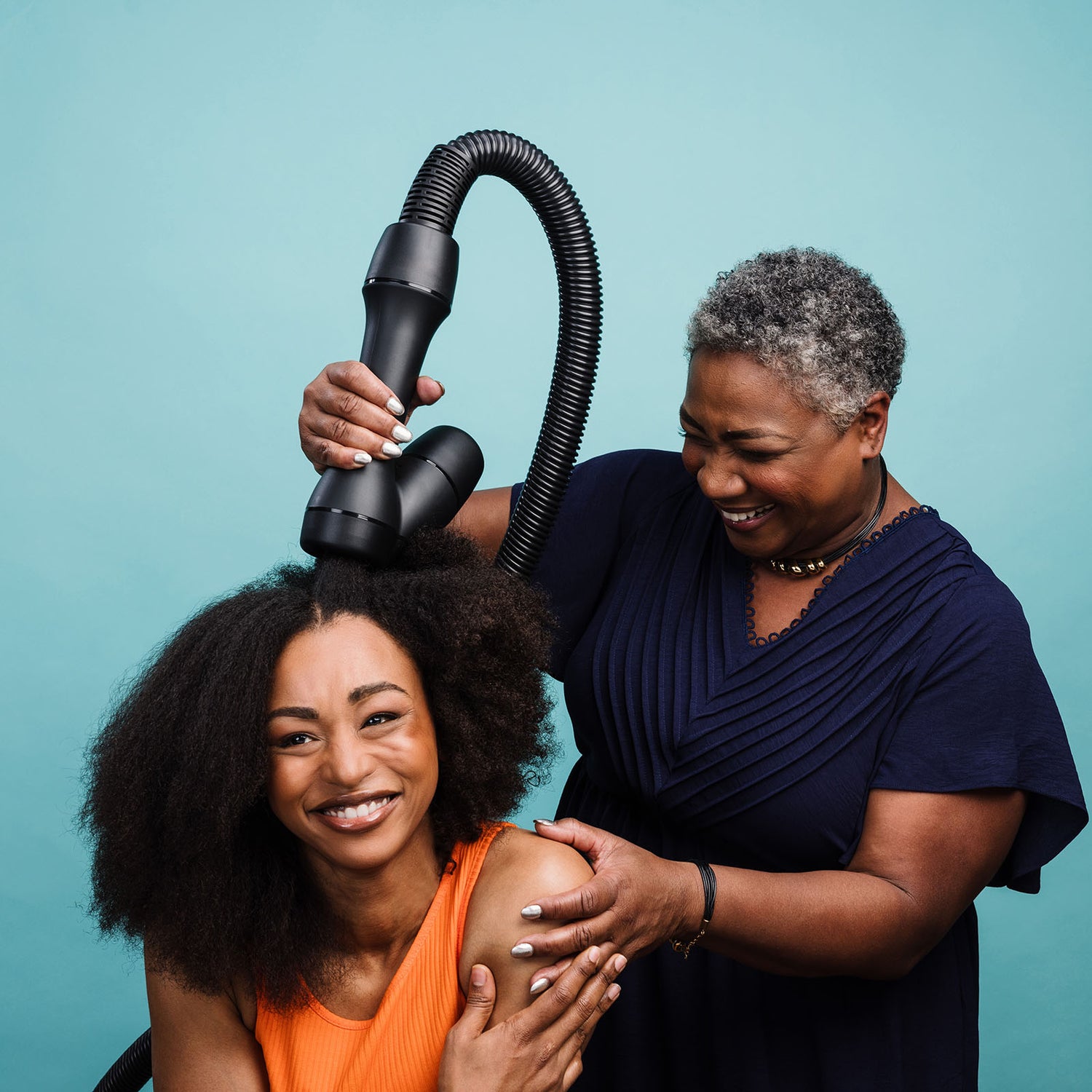 Older woman with short gray hair uses the RevAir dryer on a younger, seated woman's coily hair. Both are smiling and laughing.