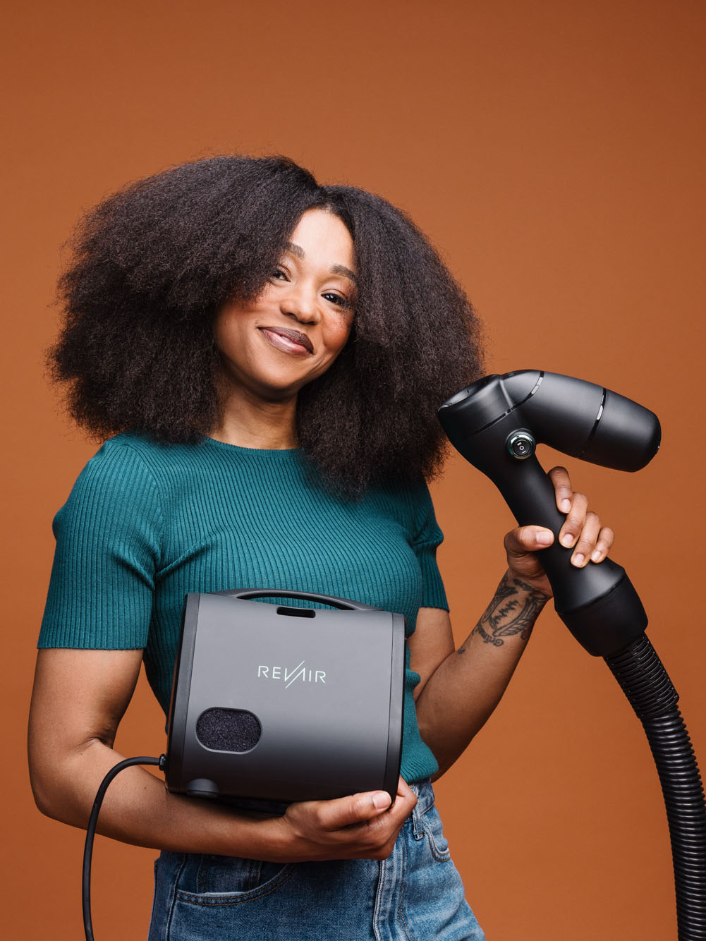 Woman with coily hair smiles and holds up the RevAir Reverse-Air Dryer and wand