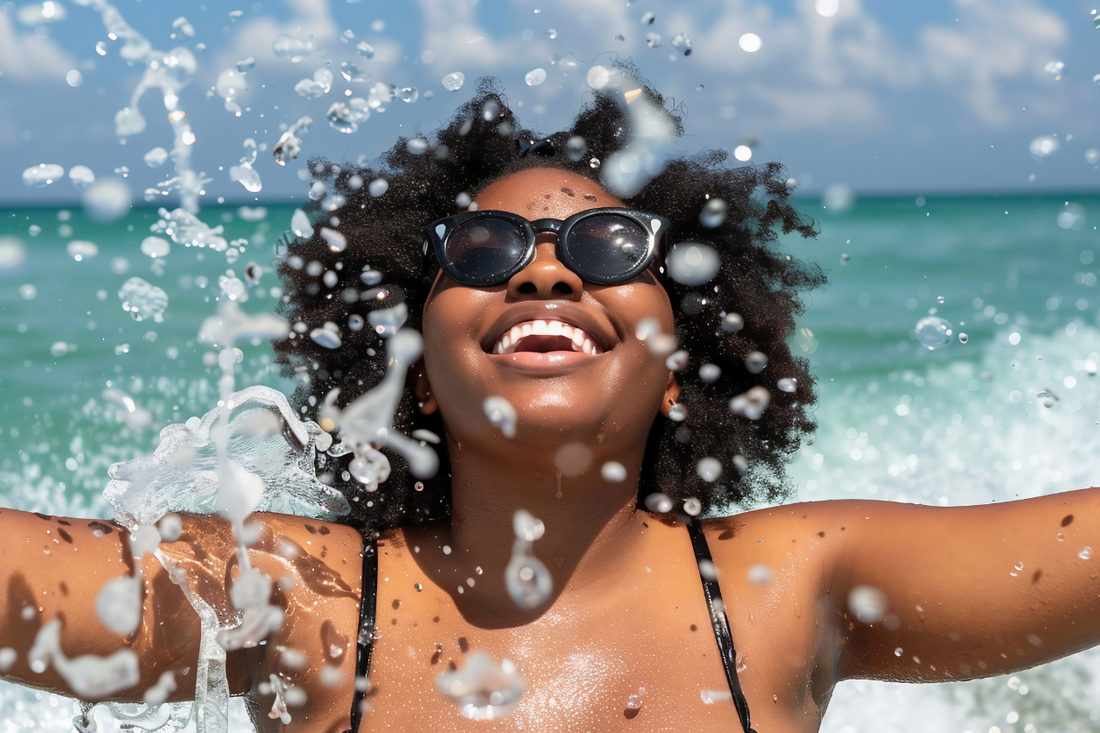 Joyful woman with natural afro hair and sunglasses enjoying ocean waves — celebrating carefree summer fun while embracing textured hair in beach environments.