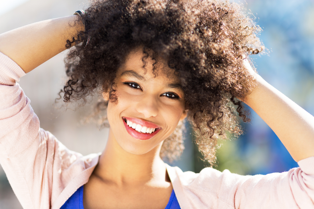 Smiling woman with natural curly hair, lifting her curls with both hands, standing outdoors in soft natural light.