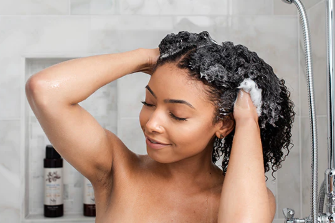 Woman with curly hair washing her hair in the shower, eyes closed, gently massaging shampoo into her curls with both hands.