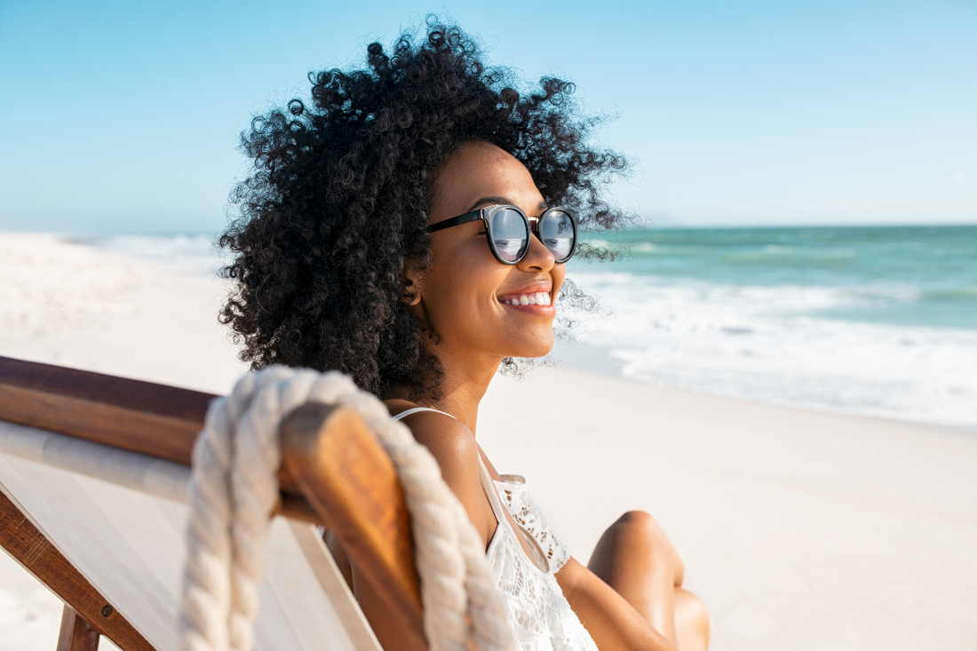 Smiling woman with natural curly hair wearing sunglasses, relaxing on a lounge chair at the beach with the ocean in the background on a bright sunny day.