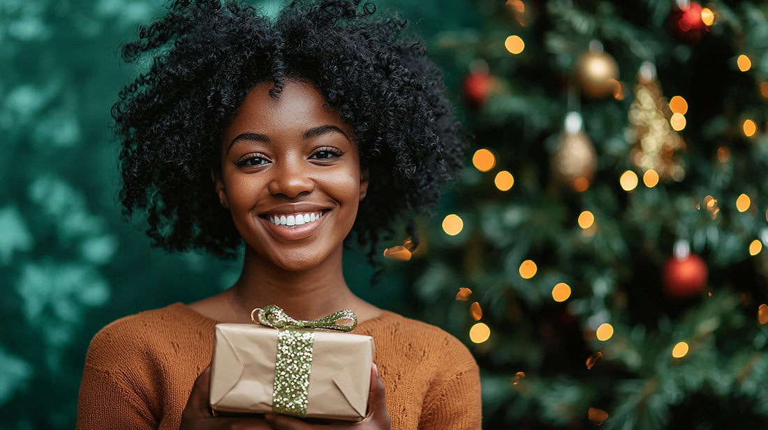 Smiling woman with natural curly hair holding a wrapped gift in front of a softly lit Christmas tree, conveying warmth, joy, and holiday celebration.