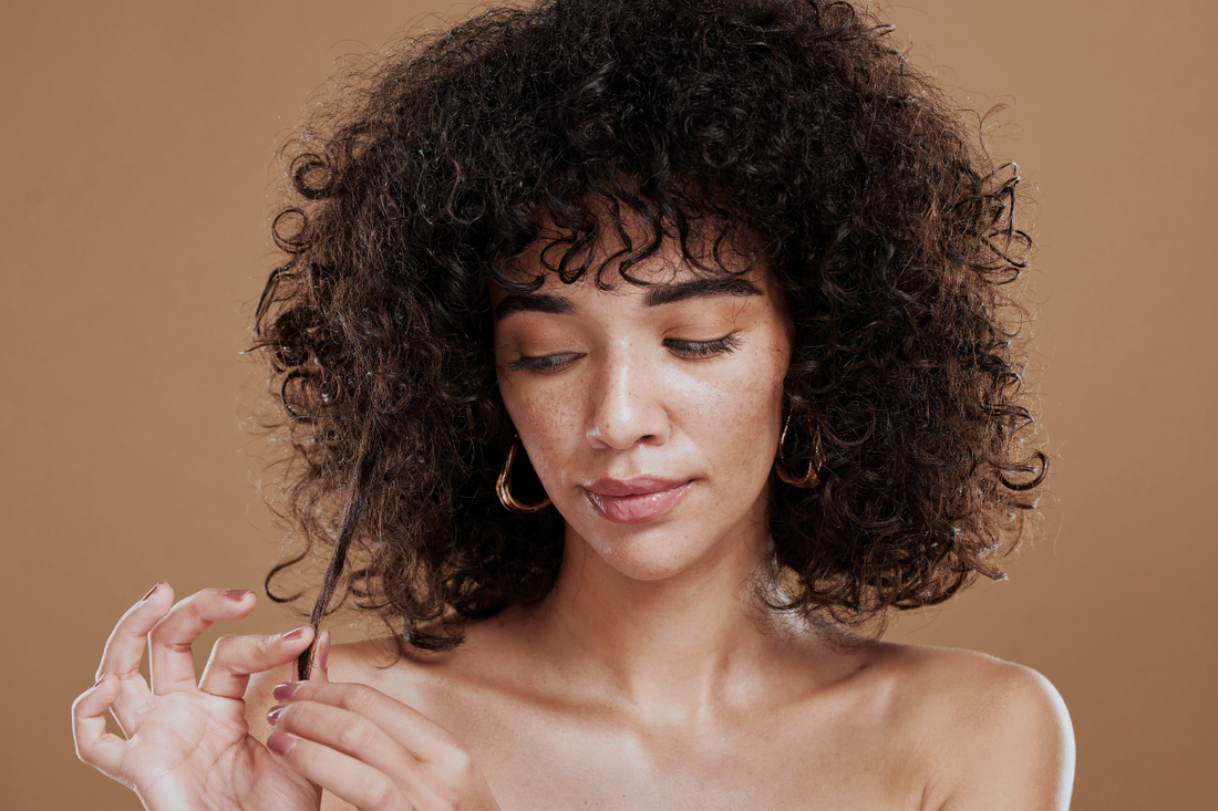 Woman with voluminous curly hair examining a strand held between her fingertips, against a warm tan studio background.