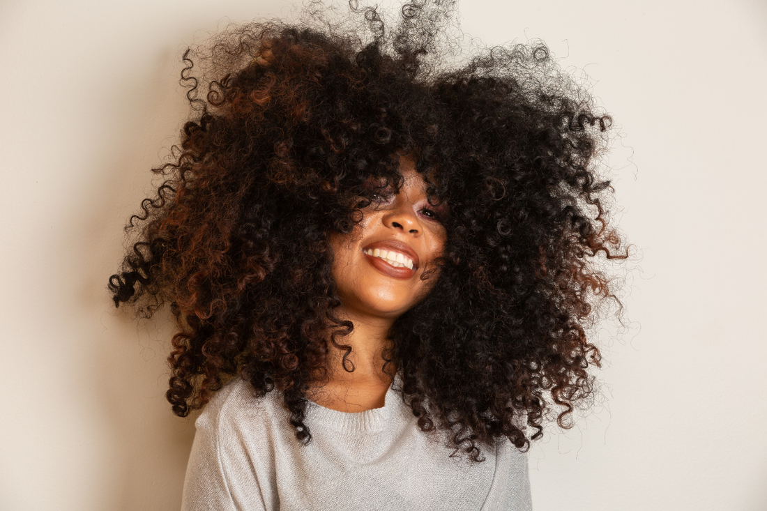 Woman with natural Type 3c/4a curls, eyes closed and smiling softly, showcasing soft volume and curl definition against a warm beige background.
