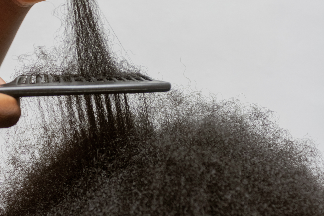 Close-up of a wide-tooth comb detangling a section of thick, coily natural hair, with several loose strands visible caught in the comb.
