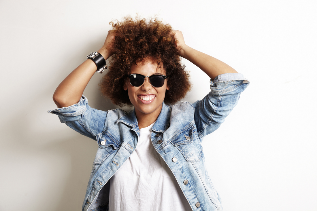 Smiling woman with natural curly hair wearing sunglasses and a denim jacket, holding her hair up against a white background. Frizzy hair texture shown in high humidity.