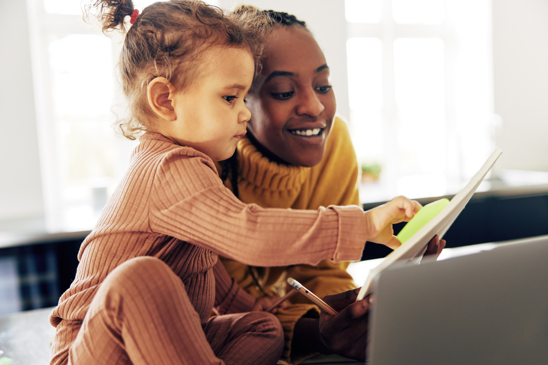 Smiling woman working with a toddler on her lap, both looking at a tablet together in a bright home setting.