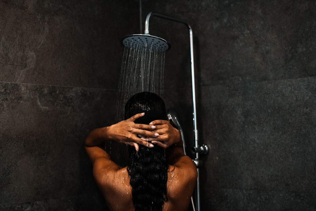 Woman washing her long dark hair under a rainfall showerhead, seen from behind on wash day