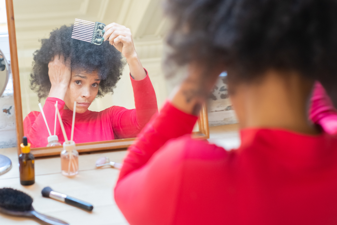 A Black woman in a red top combing her natural hair while looking in a mirror, with hair care products arranged on the vanity in front of her.