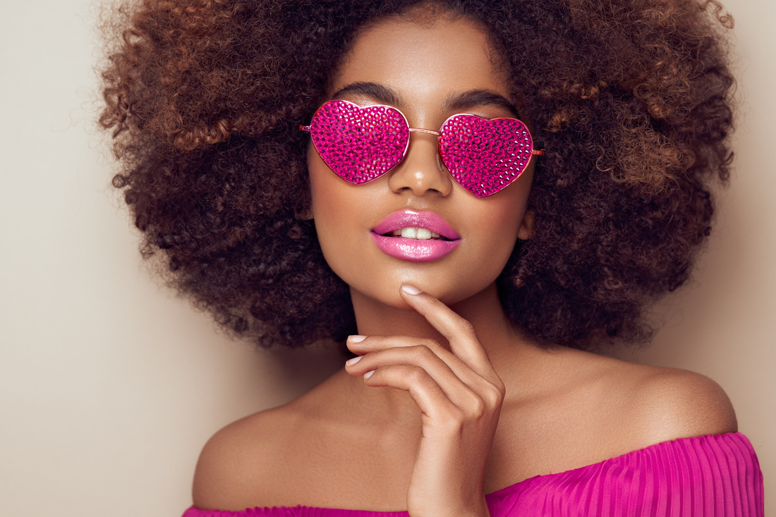 Woman with voluminous natural curls wearing pink heart-shaped sunglasses, celebrating hair self-love and Curlentine’s confidence