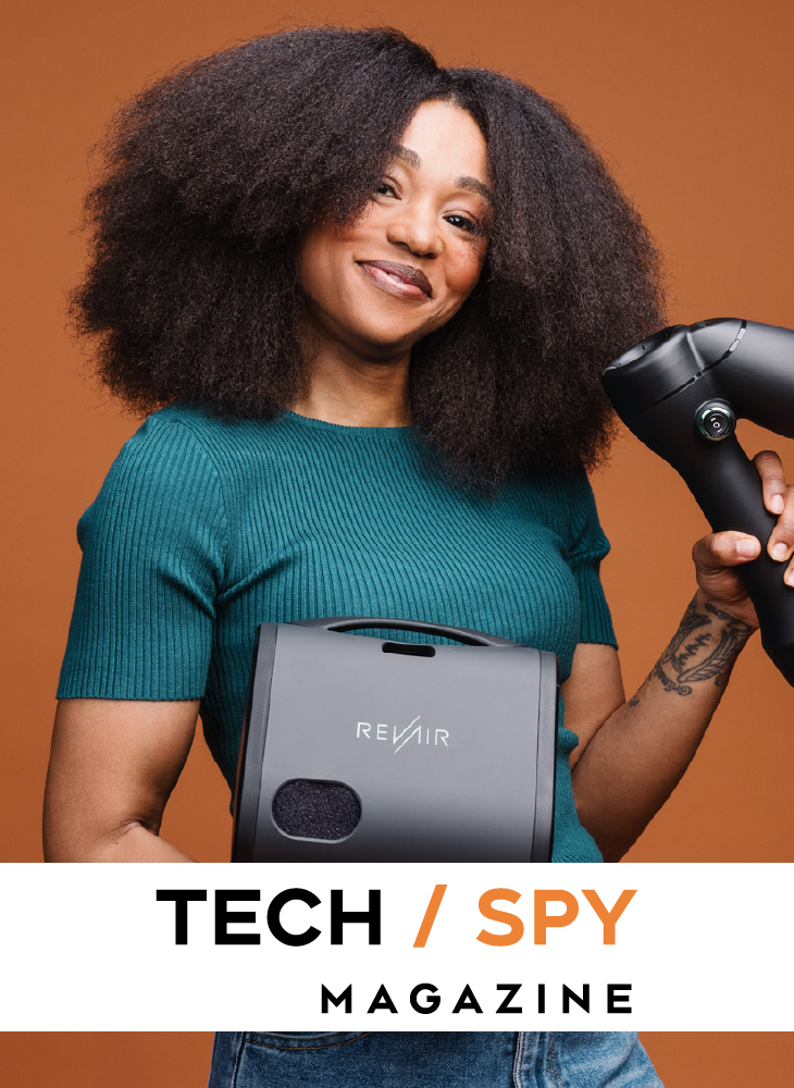A smiling Black woman with a large natural afro holding the RevAir Reverse-Air Dryer unit and wand against an orange background, with the Tech Spy Magazine logo displayed below