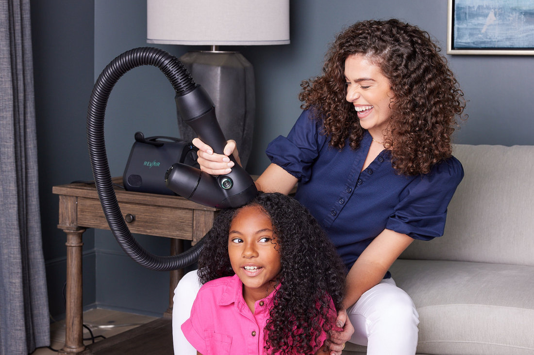 Smiling mother using a RevAir Reverse-Air Dryer on her daughter’s curly hair in a living room setting, showcasing a gentle and effective at-home styling routine.