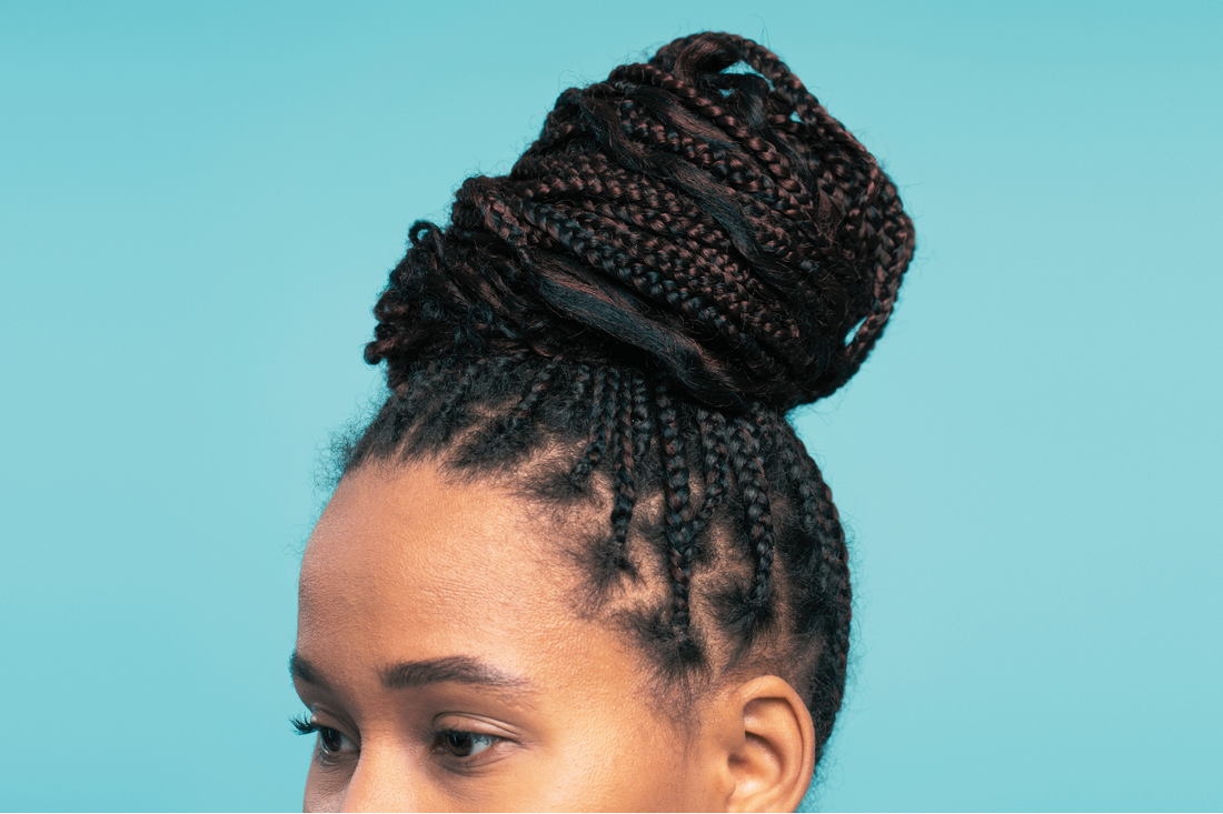 Close-up of a woman with neatly styled micro braids gathered into a high bun, showcasing a protective hairstyle against a solid light blue background.