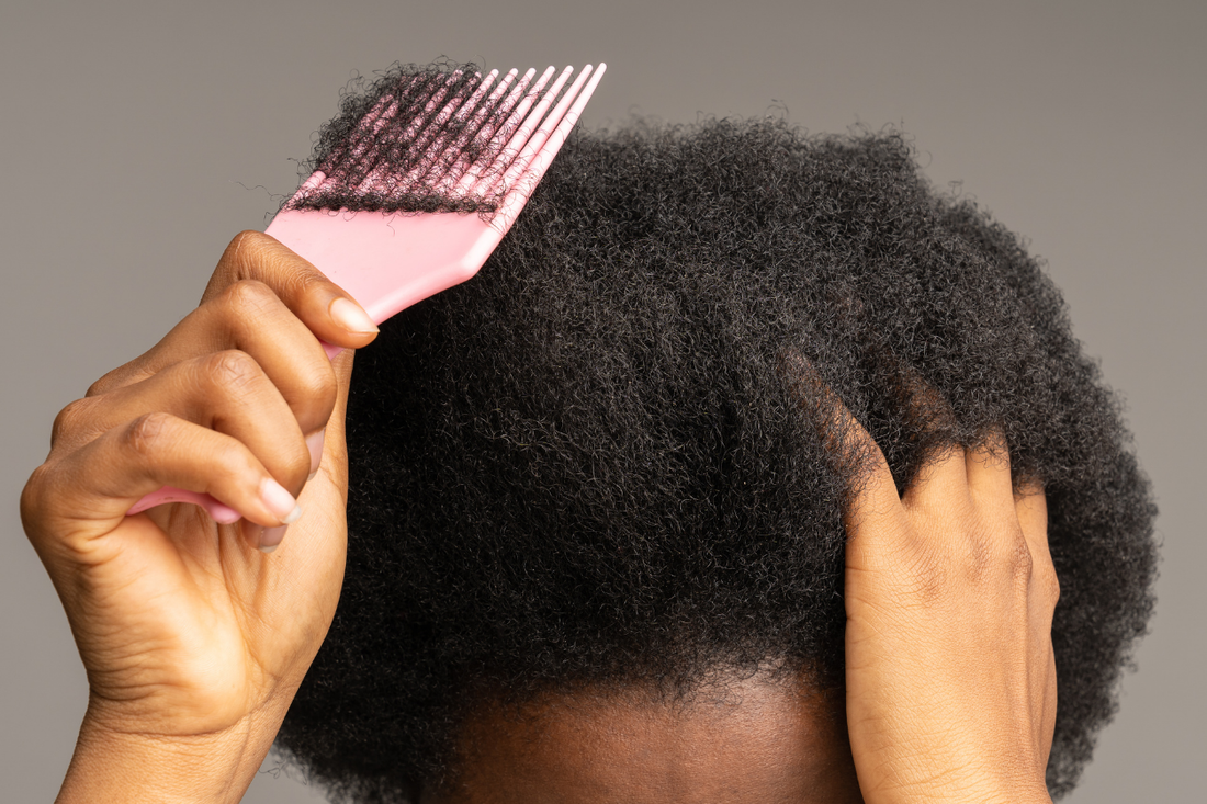 Woman using a pink wide-tooth comb to detangle a section of thick, coily type 4 hair, with her free hand steadying the roots.