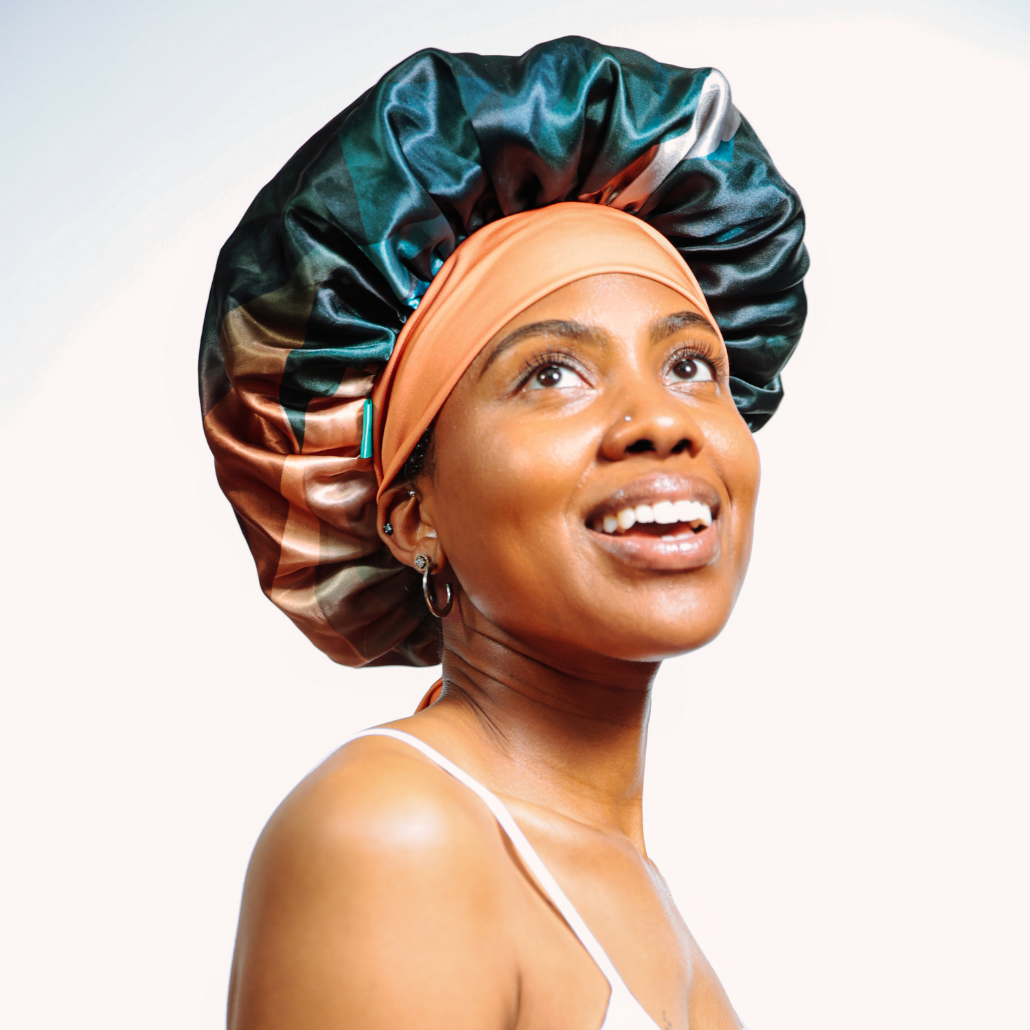 A smiling woman looking upward while wearing a colorful satin bonnet in shades of green, copper, and beige, paired with an orange head wrap. She is dressed in a white spaghetti strap top against a plain white background.