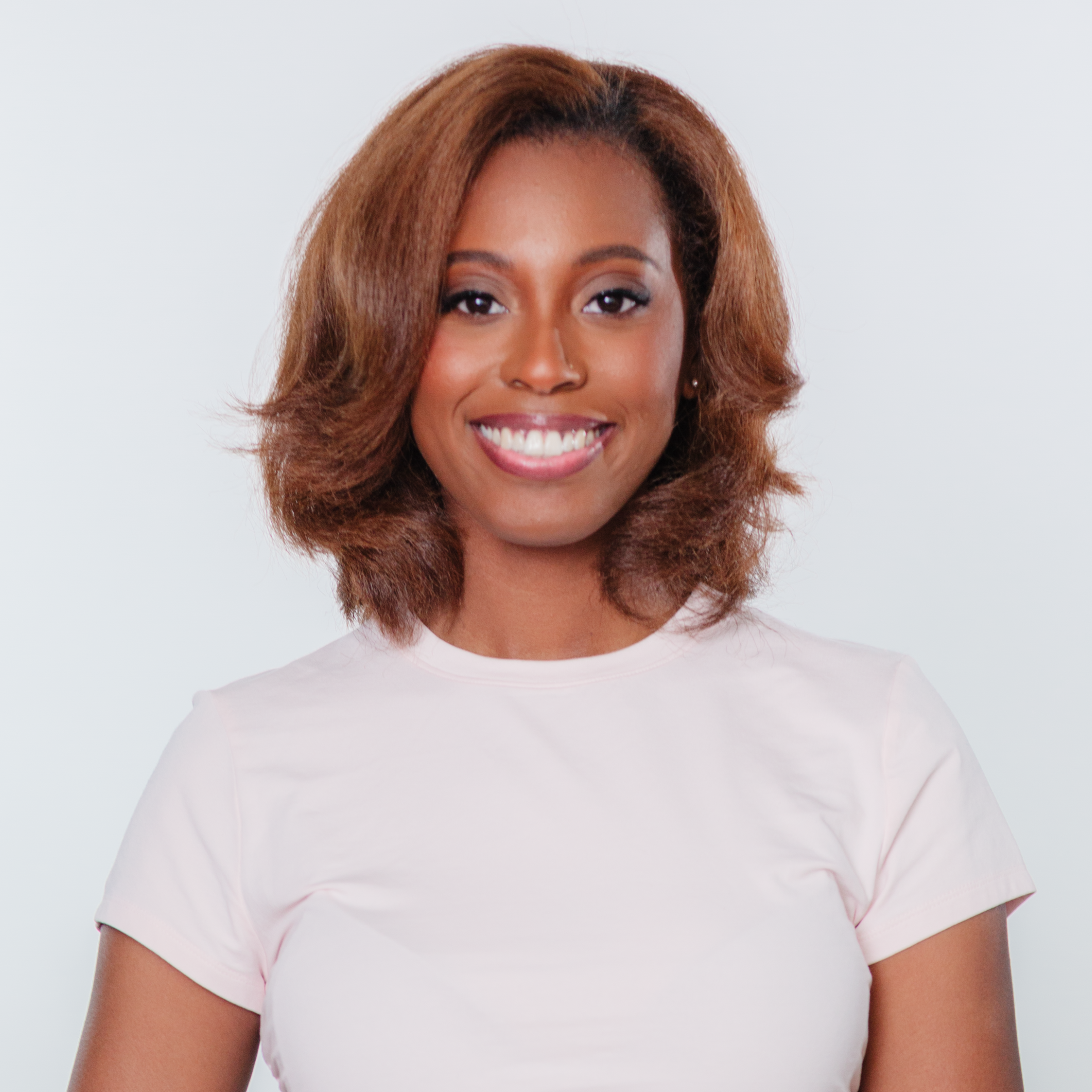 Smiling woman with shoulder-length smooth blown-out hair styled with soft layers and flipped ends, wearing a light pink short-sleeve top against a light gray background. Her hair has a voluminous, silky finish with warm caramel-brown tones.